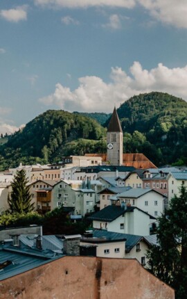 Stadtansicht von Hallein, umgeben von grünen Hügeln und einer Kirche mit Uhrturm im Zentrum unter blauem Himmel.