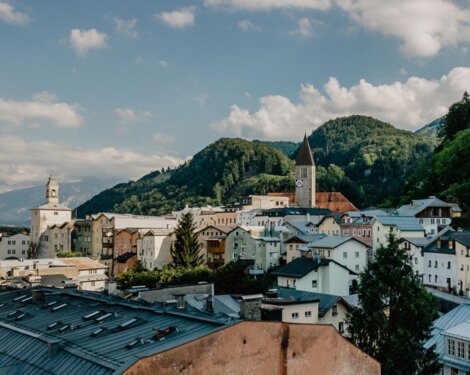 A picturesque townscape in Hallein, Austria, with colorful buildings, a church with a clock tower, and forested hills under a partly cloudy sky.
