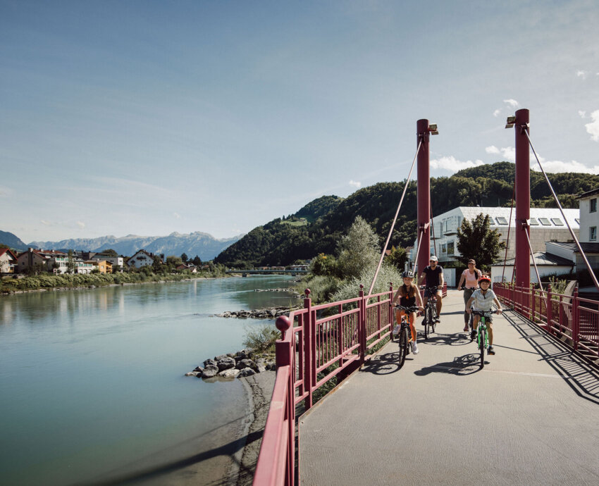 Familie radelt über eine rote Fußgängerbrücke an der Salzach in Hallein, Berge im Hintergrund.