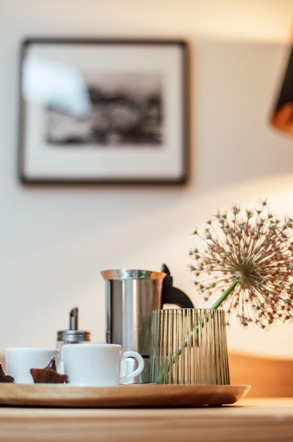 Tablett mit Kaffeetassen, Zuckerdose und dekorativer Blume auf einem Tisch, im Hintergrund eine Lampe und ein Bild an der Wand.