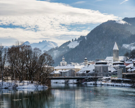 Winterliche Ansicht von Hallein: verschneite Dächer, Kirchturm und Brücke an der Salzach vor schneebedeckten Bergen.