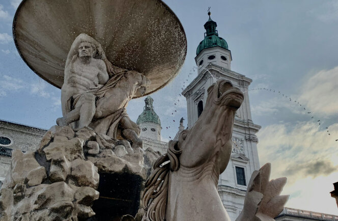 Stone fountain with mythological figures and horses in a historic square, under a cloudy sky. Tower with green dome in the background.