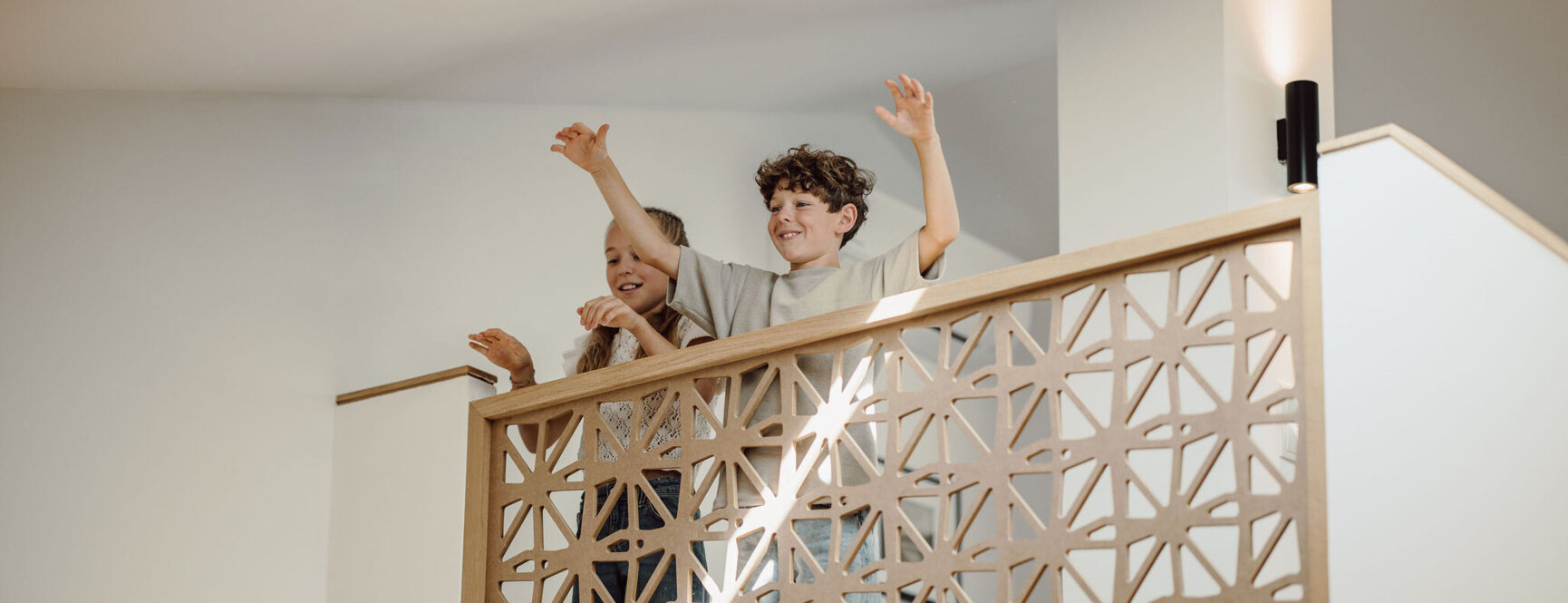 Zwei Kinder winken fröhlich von einer Galerie mit dekorativem Holzgeländer in einem hellen, modernen Innenraum.